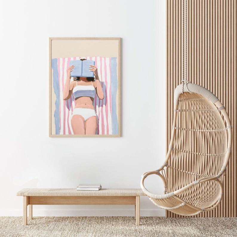 May include: A framed illustration of a person lying on a striped beach towel, reading a book. The person is wearing a white bikini and has their face obscured by the book. The illustration is in a minimalist style with muted colors.