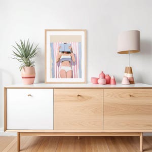 May include: A framed illustration of a person laying on a striped beach towel and reading a book. The person is wearing a white bikini and has their hair pulled back. The illustration is in a light, airy style with pink and blue accents.