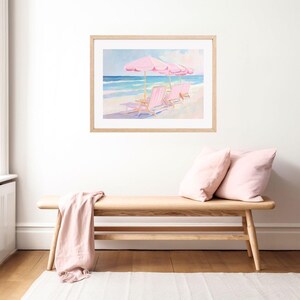 May include: A framed painting of a beach scene with three pink lounge chairs and pink umbrellas. The chairs are facing the ocean, which is a light blue color. The sky is a light blue with some clouds. The painting is in a light wood frame.