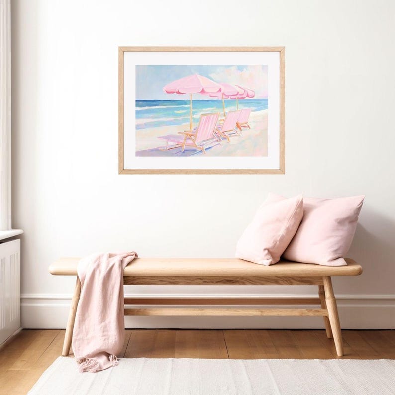 May include: A framed painting of a beach scene with three pink lounge chairs and pink umbrellas. The chairs are facing the ocean, which is a light blue color. The sky is a light blue with some clouds. The painting is in a light wood frame.
