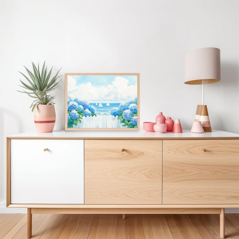 May include: A framed art print depicts a coastal scene with blue hydrangeas, a white picket fence, and sailboats on the water under a blue sky. The artwork is displayed on a white and wood-toned cabinet, alongside a potted plant and decorative pink vases.