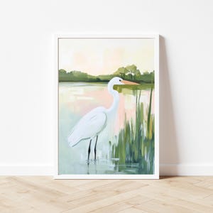 K&ouml;nnte beinhalten: Gerahmter Kunstdruck eines Graureihers im Wasser. Das Gem&auml;lde zeigt eine ruhige Landschaft mit sanften Rosa- und Gr&uuml;nt&ouml;nen, Schilf und einem verschwommenen Hintergrund von B&auml;umen. Der Vogel hat einen langen Hals und einen orangefarbenen Schnabel.