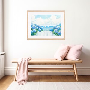 May include: A framed watercolor print depicts a coastal scene with blue hydrangeas, a white picket fence, and sailboats on the horizon. The artwork is displayed above a wooden bench with pink pillows and a draped pink throw, creating a serene atmosphere.