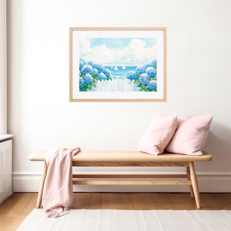 May include: A framed watercolor print depicts a coastal scene with blue hydrangeas, a white picket fence, and sailboats on the horizon. The artwork is displayed above a wooden bench with pink pillows and a draped pink throw, creating a serene atmosphere.
