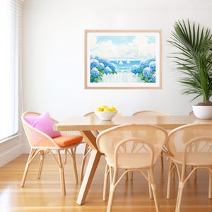 May include: A framed watercolor painting depicts a coastal scene with blue hydrangeas, a white picket fence, and sailboats on the ocean. The painting hangs above a wooden dining table with woven chairs, a pink pillow, and a bowl of lemons.