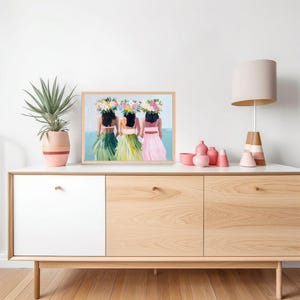 Op de afbeelding: Een ingelijst schilderij van drie vrouwen die bloemenkransen en kleurrijke rokken dragen. De vrouwen staan voor een blauwe en witte achtergrond. Het schilderij staat op een houten plank met een lamp en vazen.