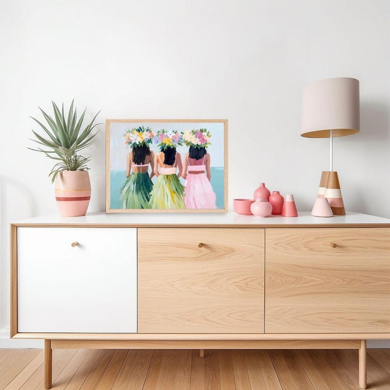 Op de afbeelding: Een ingelijst schilderij van drie vrouwen die bloemenkransen en kleurrijke rokken dragen. De vrouwen staan voor een blauwe en witte achtergrond. Het schilderij staat op een houten plank met een lamp en vazen.