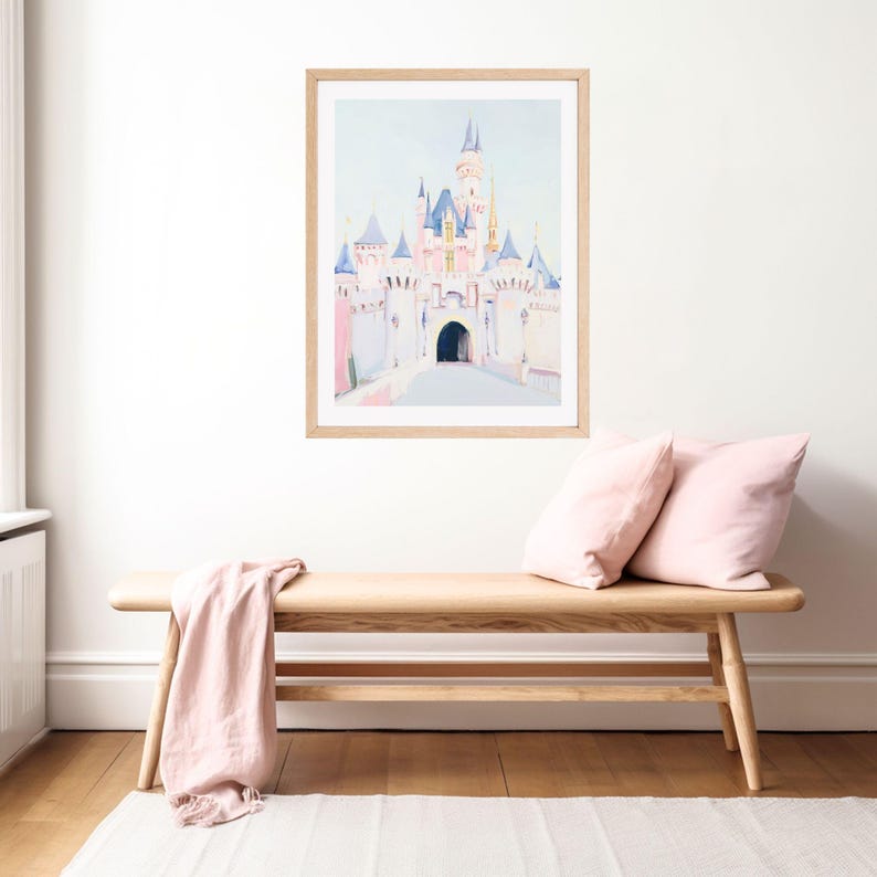 May include: Watercolor painting of a fairytale castle in a wooden frame. The castle features pink and blue turrets and spires. The artwork is displayed above a wooden bench with pink pillows and a pink throw.