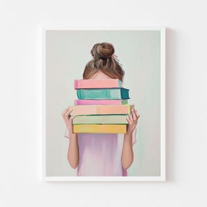 May include: A framed art print featuring a person holding a stack of colorful books. The books are in shades of pink, blue, green, and yellow. The person is wearing a light pink shirt and has their hair up in a bun.