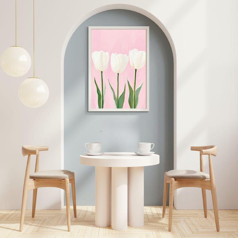 May include: Three white tulips with green stems on a pink background in a white frame. The frame is hanging on a light blue wall. There is a round white table with two wooden chairs in front of the wall.