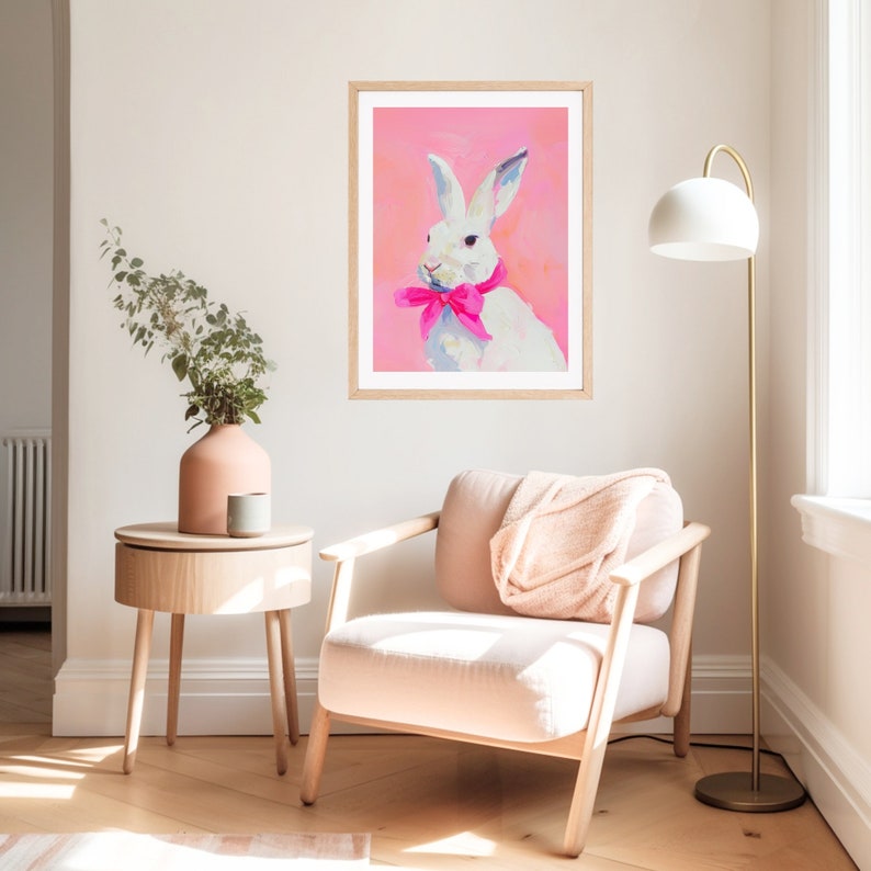 May include: A white rabbit with a pink bow tie is painted on a pink background. The painting is framed and hanging on a white wall in a room with a chair and a side table.