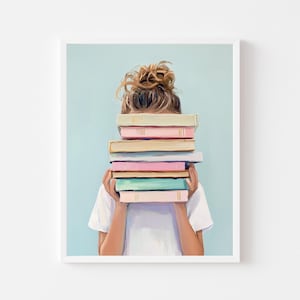 May include: A framed print featuring a person holding a stack of colorful books in front of their face. The books have pastel-colored spines, and the background is a light blue. The person is wearing a white t-shirt.