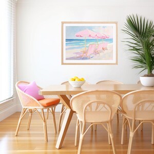 May include: A watercolor painting of pink beach chairs and umbrellas on a white sandy beach with blue ocean waves in the background. The painting is framed and hanging on a white wall in a dining room with a wooden table and wicker chairs.