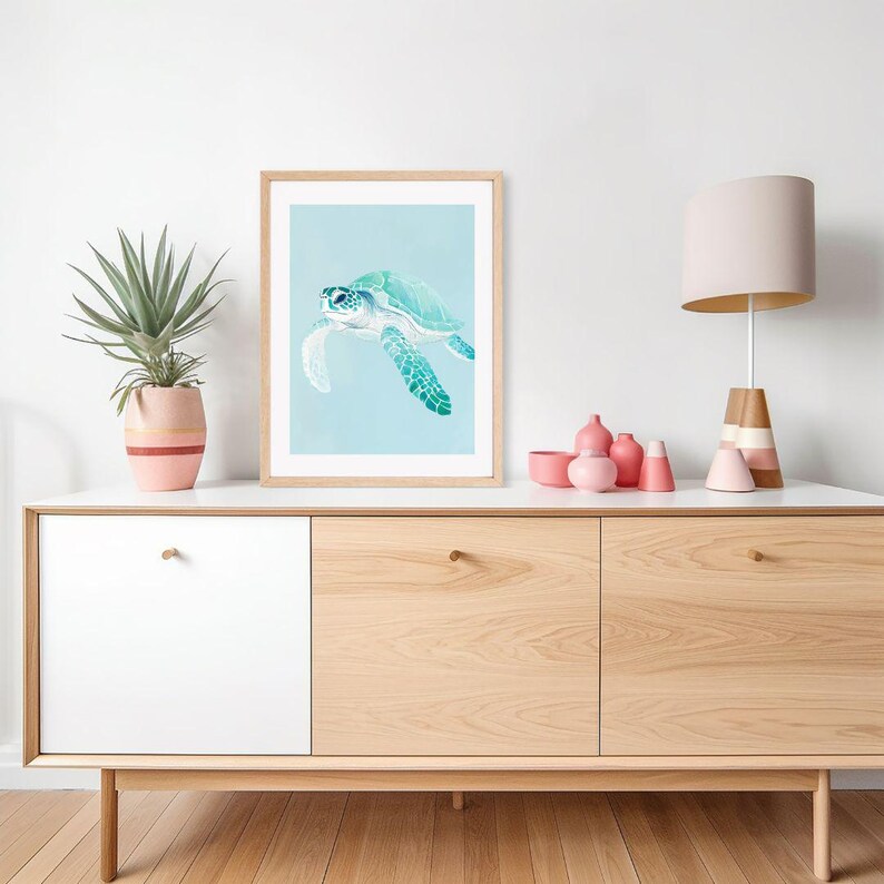 Puede incluir: Una impresi&oacute;n enmarcada de una pintura de acuarela de una tortuga marina nadando en agua azul. La impresi&oacute;n est&aacute; colgada en una pared blanca sobre un gabinete de madera clara con tres cajones.