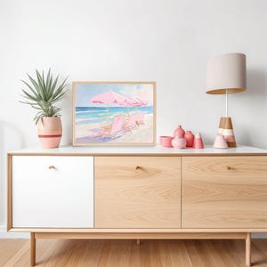 May include: A framed watercolor painting of pink beach chairs under pink umbrellas on a white sandy beach with blue ocean waves. The painting is hanging on a light wood cabinet with two white doors and two light wood doors.