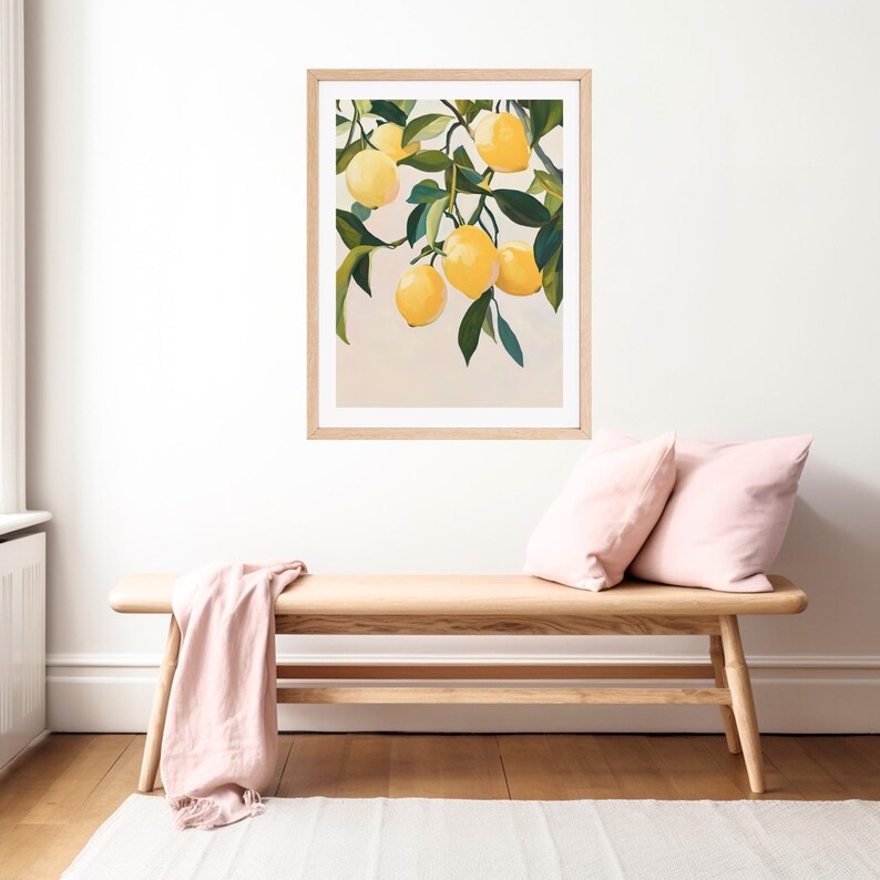 May include: A framed art print featuring a still life of lemons on a branch with green leaves. The lemons are yellow, and the leaves are green. The artwork is in a light wood frame, displayed above a wooden bench with pink pillows and a pink throw.