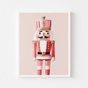 May include: A framed print of a pink and red nutcracker soldier. The nutcracker has a black mustache, white hair, and a gold-colored hat and epaulets. The background is a solid light pink color.