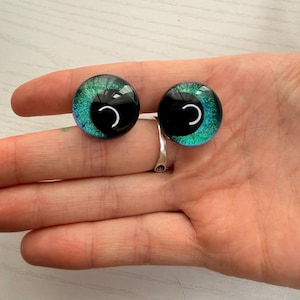May include: Two glass doll eyes with a black pupil and a teal and green iris. The eyes are round and have a reflective surface. The eyes are held in a person's hand.