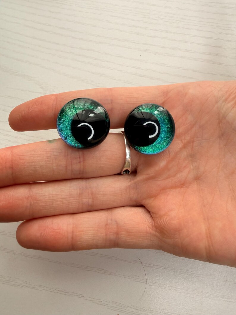May include: Two glass doll eyes with a black pupil and a teal and green iris. The eyes are round and have a reflective surface. The eyes are held in a person's hand.
