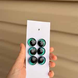 May include: Six glass eye cabochons in shades of teal and black are displayed on a white card. The eyes have a glossy finish and a detailed iris design. The card is held against a light brown background.