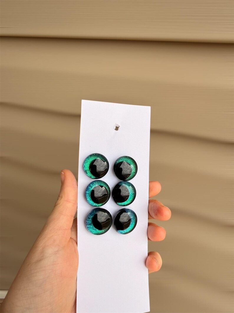May include: Six glass eye cabochons in shades of teal and black are displayed on a white card. The eyes have a glossy finish and a detailed iris design. The card is held against a light brown background.