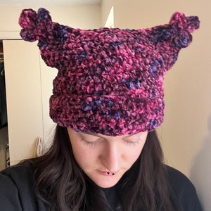 May include: A purple and pink crocheted beanie with two fluffy ear flaps. The beanie is made with a chunky yarn and has a textured look.