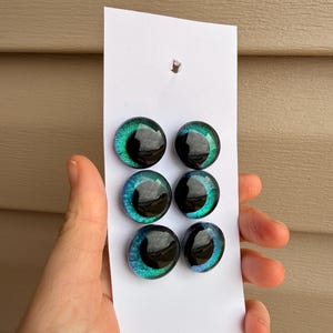May include: Six iridescent glass eye cabochons in shades of green and blue, with a black pupil. The cabochons are arranged on a white card. The eyes are approximately 0.75 inches in diameter.