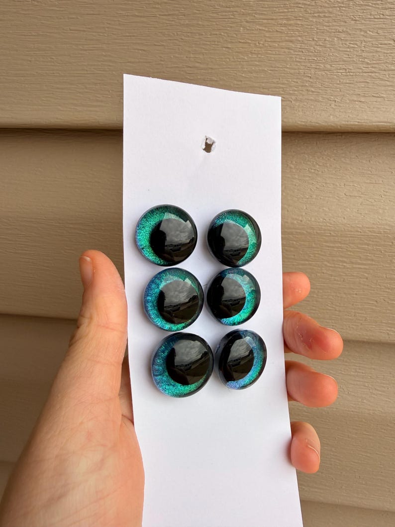 May include: Six iridescent glass eye cabochons in shades of green and blue, with a black pupil. The cabochons are arranged on a white card. The eyes are approximately 0.75 inches in diameter.