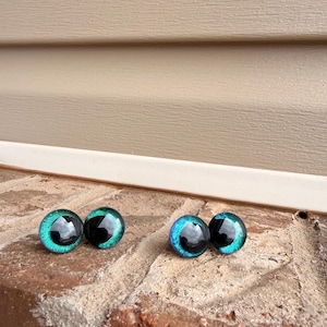 May include: Four glass doll eyes with a black pupil and a teal and blue iris. The eyes are round and have a reflective surface. They are displayed on a brick surface.