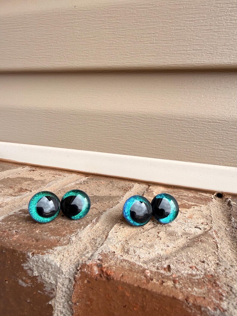 May include: Four glass doll eyes with a black pupil and a teal and blue iris. The eyes are round and have a reflective surface. They are displayed on a brick surface.