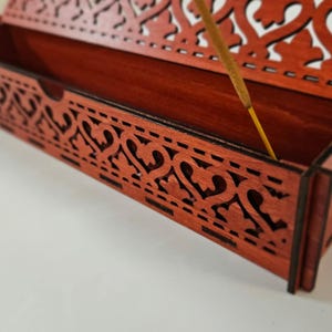 May include: A rectangular wooden incense burner box with a reddish-brown finish and intricate laser-cut heart and leaf patterns along the sides and lid. A single thin incense stick is positioned upright inside the open wooden container.