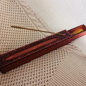 Peut inclure: Une boîte à encens rectangulaire en bois avec une finition brun rougeâtre foncé et des motifs floraux complexes découpés au laser sur le couvercle. La boîte dispose d'un compartiment coulissant contenant des bâtonnets d'encens jaunes.