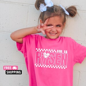 May include: A pink t-shirt with a white graphic that says "He is Risen" with a checkered pattern and a heart with a cross inside.
