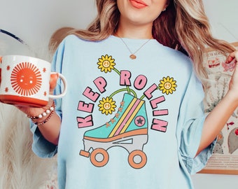 May include: Light blue t-shirt with a retro roller skate graphic. The graphic features a teal skate with pink and yellow accents, and the words "Keep Rollin" in pink. The shirt also has a graphic of a smiling sun on a mug.