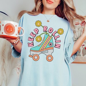 May include: Light blue t-shirt with a retro roller skate graphic. The graphic features a teal skate with pink and yellow accents, and the words "Keep Rollin" in pink. The shirt also has a graphic of a smiling sun on a mug.