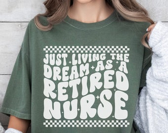 May include: A green t-shirt with a white graphic that says "Just Living the Dream as a Retired Nurse" in a retro font. The graphic is surrounded by a checkered border.