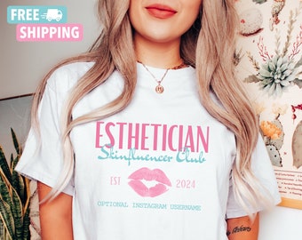 May include: White t-shirt with the text "Esthetician Skinfluencer Club" in pink and blue lettering. A pink lip print is centered below the text with "Est 2024" on either side. The text "Optional Instagram Username" is below the lip print.