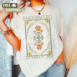 May include: A cream colored t-shirt with the text "ENCHANTED to teach you" in a vintage style font. The text is surrounded by a floral design with green leaves, orange and yellow flowers, and gold stars.