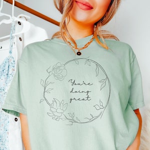 May include: A light green t-shirt with the words "You're doing great" in a floral wreath design. The shirt is a solid color and has short sleeves. The text and floral design are in black.