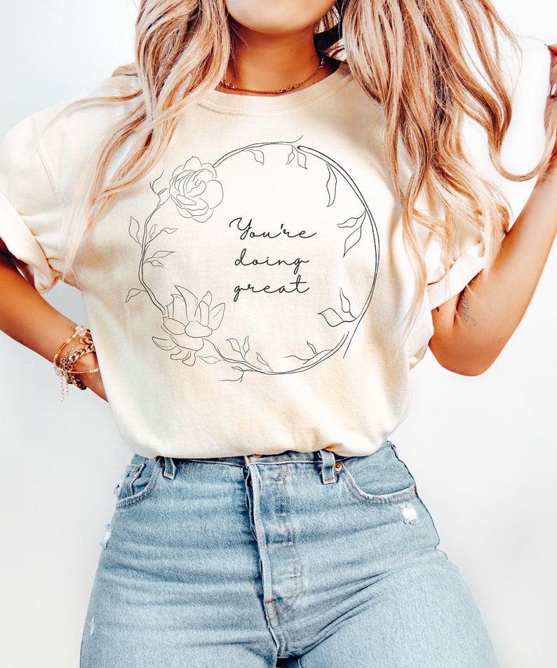May include: Cream-colored t-shirt with a floral wreath design and the words "You're doing great" in black script. The shirt is paired with light blue denim jeans. The overall aesthetic is casual and comfortable.