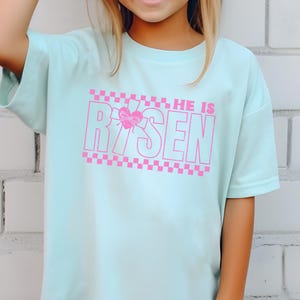 May include: A light blue t-shirt with a pink graphic that says "HE IS RISEN" with a pink checkered pattern above and below the text. A pink butterfly is in the middle of the "R" in "RISEN".