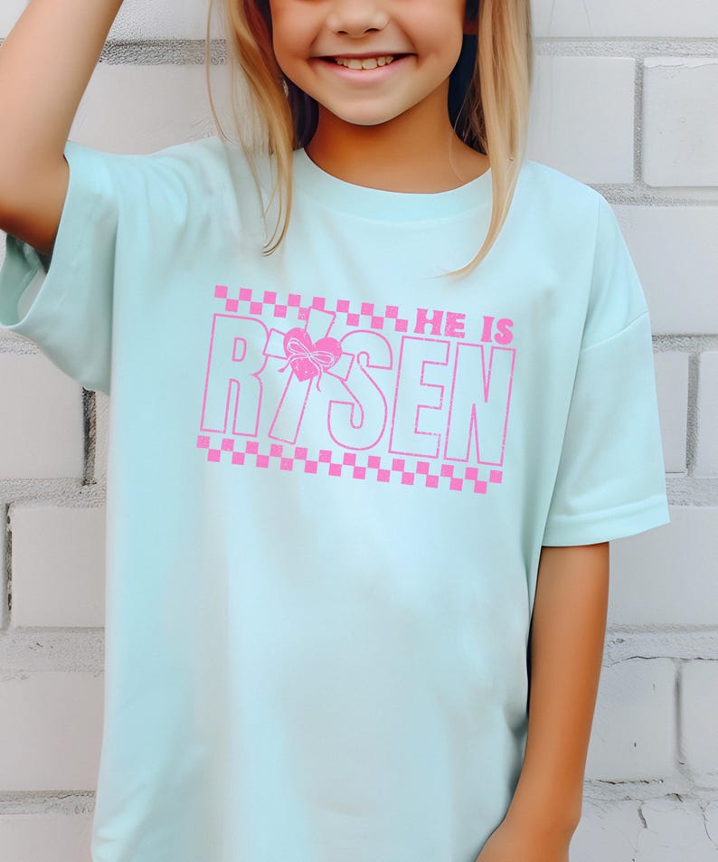 May include: A light blue t-shirt with a pink graphic that says "HE IS RISEN" with a pink checkered pattern above and below the text. A pink butterfly is in the middle of the "R" in "RISEN".