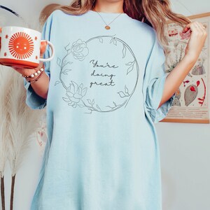 May include: Light blue t-shirt with the text "You're doing great" inside a floral wreath. A white and orange mug with a smiling sun graphic is held in the left hand.