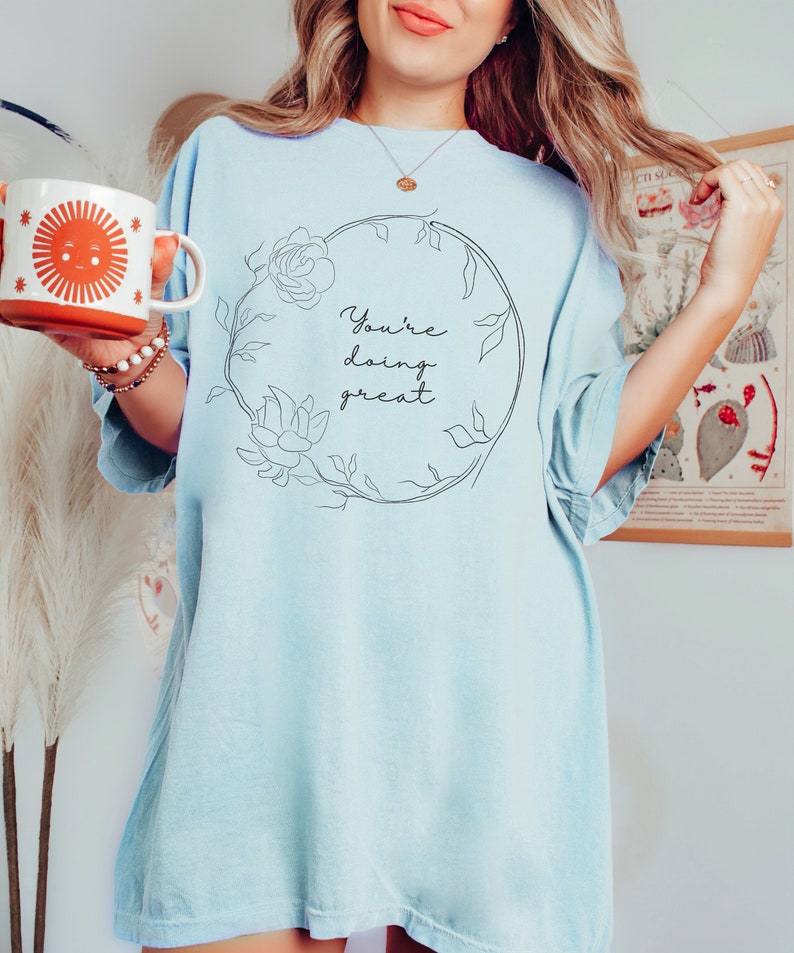 May include: Light blue t-shirt with the text "You're doing great" inside a floral wreath. A white and orange mug with a smiling sun graphic is held in the left hand.