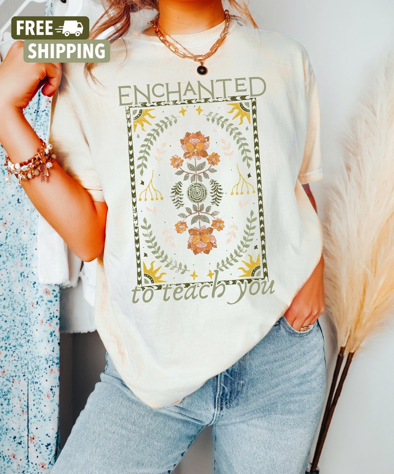 May include: A cream colored t-shirt with the text "ENCHANTED to teach you" in a vintage style font. The text is surrounded by a floral design with green leaves, orange and yellow flowers, and gold stars.