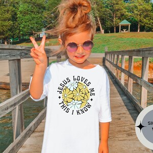 May include: A white t-shirt with a yellow and white floral design and the text "Jesus Loves Me This I Know". The shirt is shown on a young person wearing sunglasses and standing on a wooden bridge.