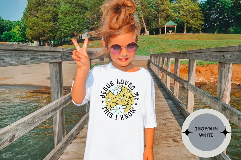 May include: A white t-shirt with a yellow and white floral design and the text "Jesus Loves Me This I Know". The shirt is shown on a young person wearing sunglasses and standing on a wooden bridge.