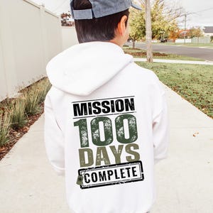 K&ouml;nnte beinhalten: Wei&szlig;er Kapuzenpullover mit dem Text "Mission 100 Days Complete" in schwarzer und gr&uuml;ner Schrift mit einem Used-Look.