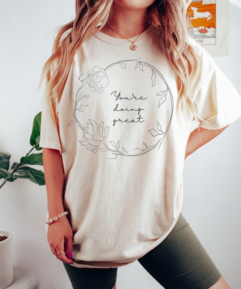 May include: Cream-colored t-shirt with the phrase "You're doing great" in a black script font, encircled by a floral wreath design. The shirt is a relaxed fit, and the overall aesthetic is casual and comfortable.
