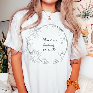 May include: White t-shirt with a floral wreath design and the text "You're doing great" in black script. The wreath features a rose and other floral elements. The shirt is a casual, comfortable style.